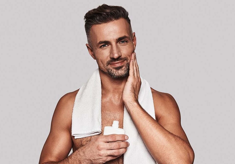 Confident in his skin. Handsome young man applying aftershave lotion and smiling while standing against grey background